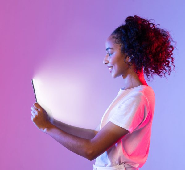 Side profile of happy black woman with curly hair, looking at her tablet whose screen illuminates her face, against purple and pink gradient neon backdrop